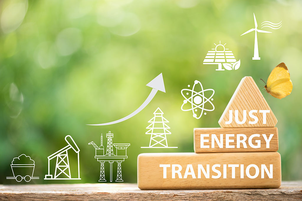 Drei gestlapelte Holzklötze mit der Aufschrift "Just Energy Transition" vor grünem Hintergrund. Links daneben der Verlauf der Energiewende dargstellt durch Grafikelemente der Energiegewinnungsarten im Verlauf der Zeit.
